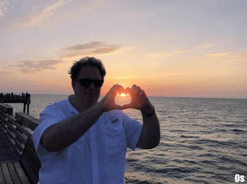 A person wearing sunglasses makes a heart shape with their hands, framing the setting sun over the ocean. The sky shows warm hues of orange and pink as the sun dips towards the horizon.