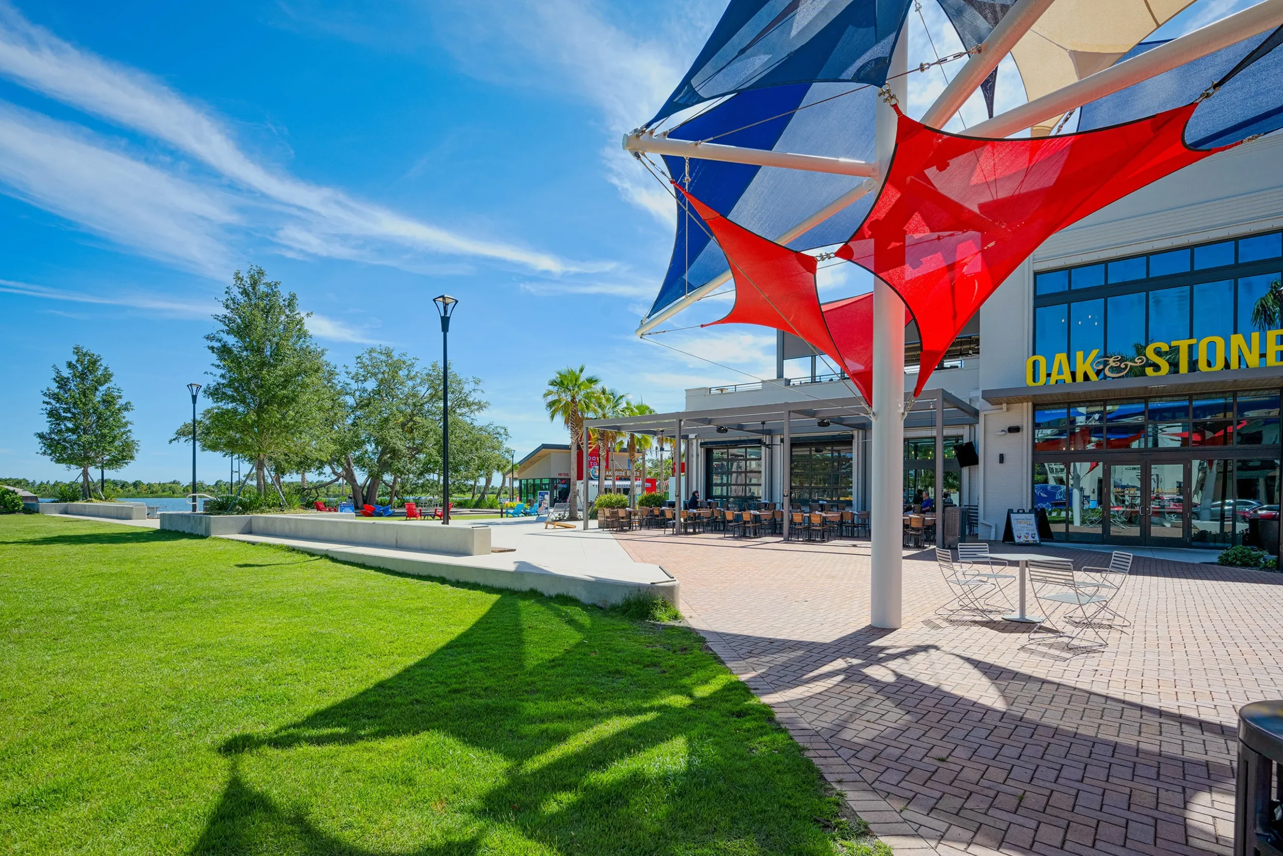 Waterfront dining area at Oak & Stone in Nokomis, FL, featuring colorful shade sails, outdoor seating, and lush green landscaping, highlighting community-oriented lifestyle and family-friendly amenities.